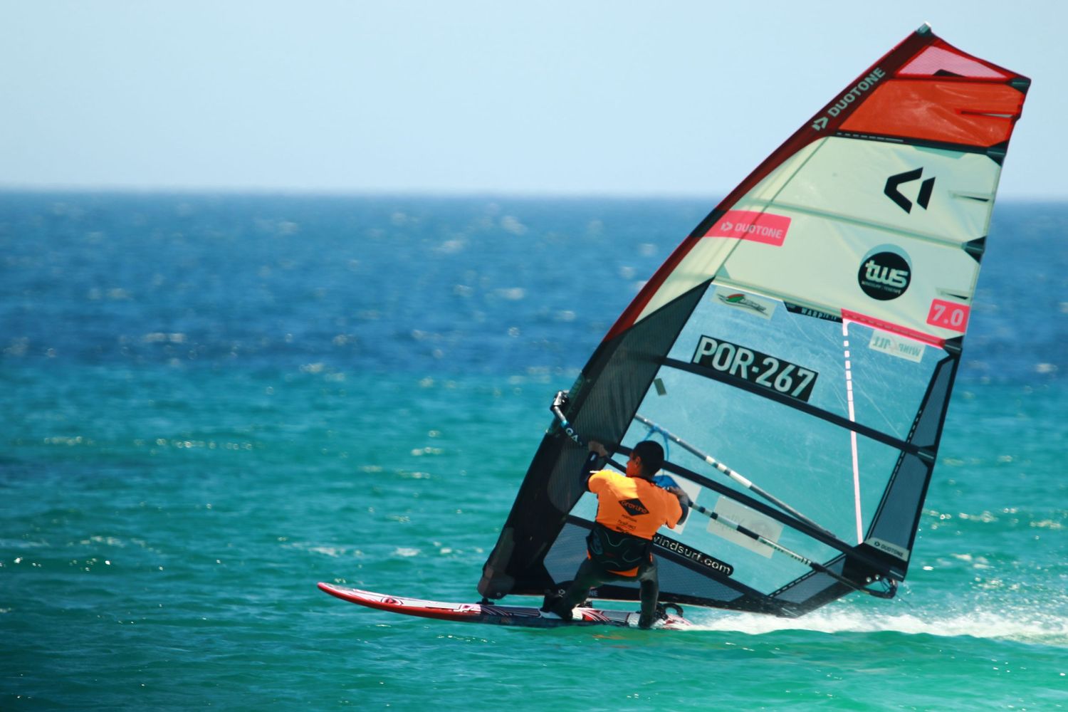 Windsurfer sailing across turquoise waters with red and white racing sail.