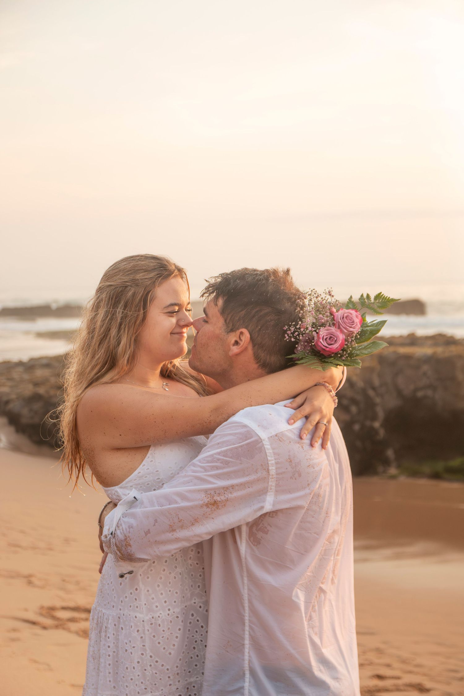 A romantic couple embraces on a sandy beach at sunset with soft golden light creating a dreamy atmosphere.