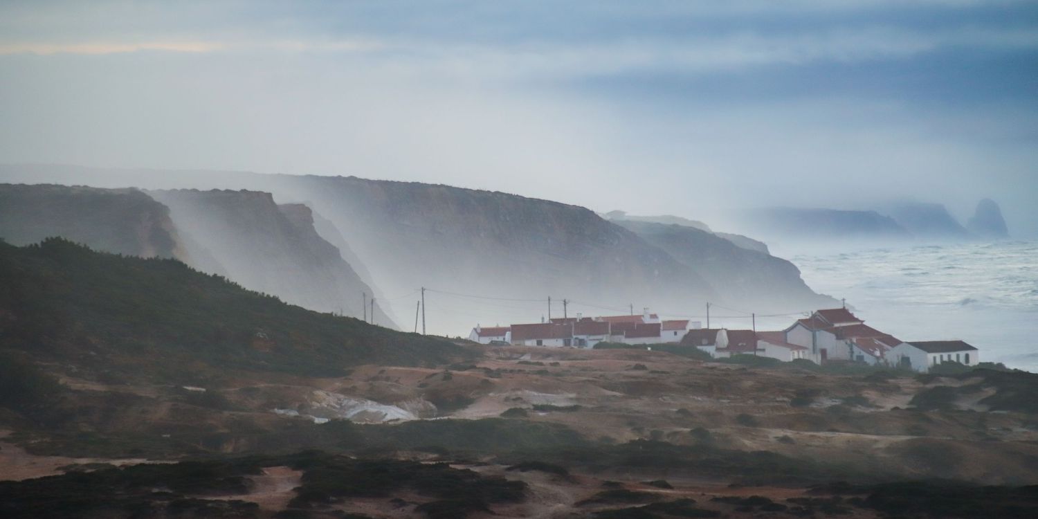 Foggy coastal landscape with buildings nestled on rolling hills near misty cliffs and ocean shore.