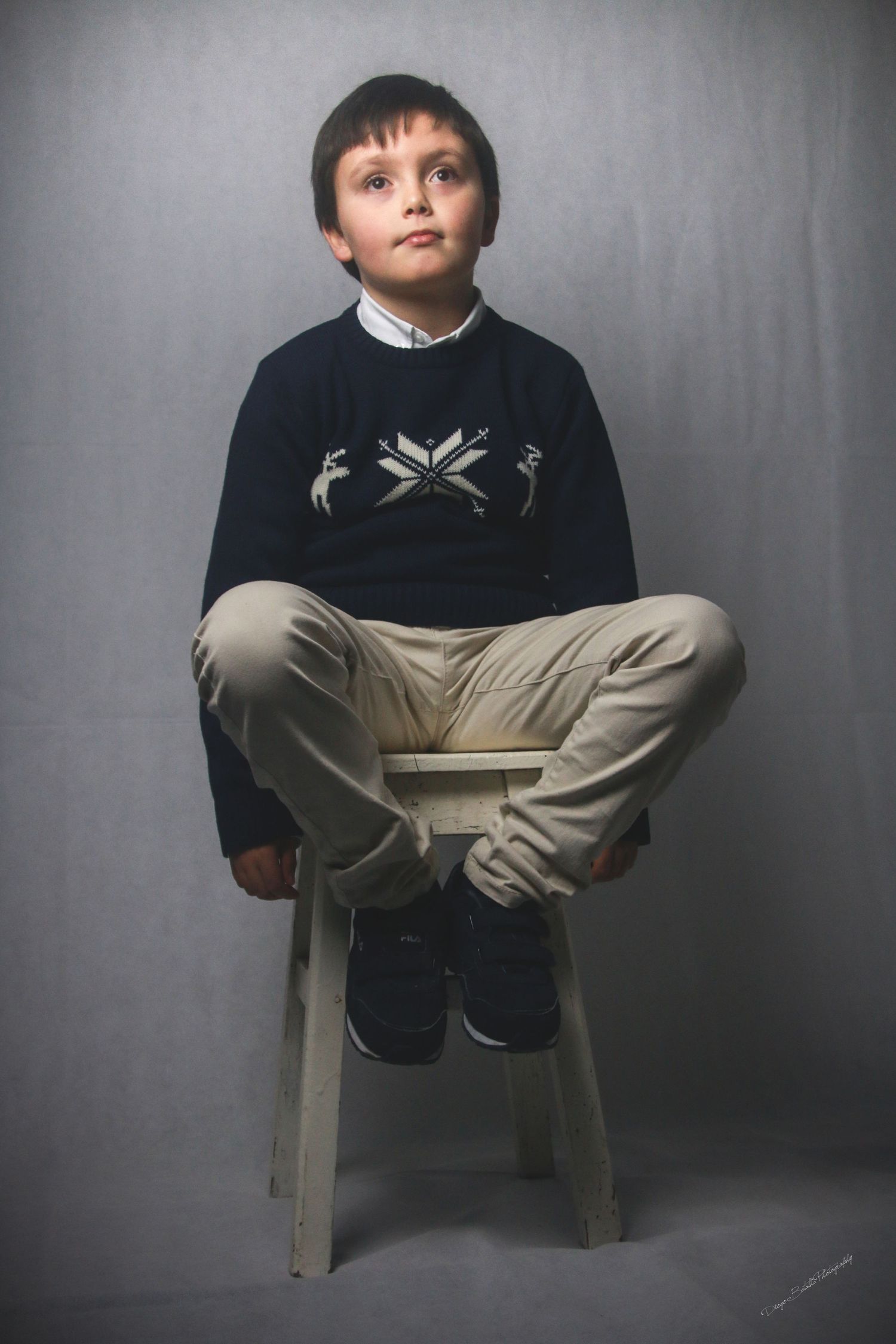 Portrait of young person in festive sweater sitting on white stool against grey backdrop.