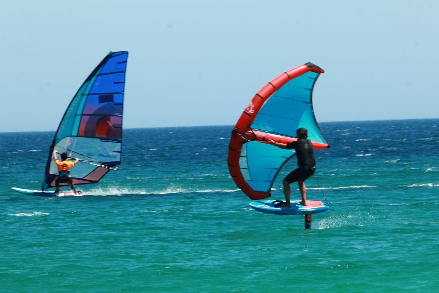 Windsurfers on foil boards with blue and red sails riding turquoise waters.