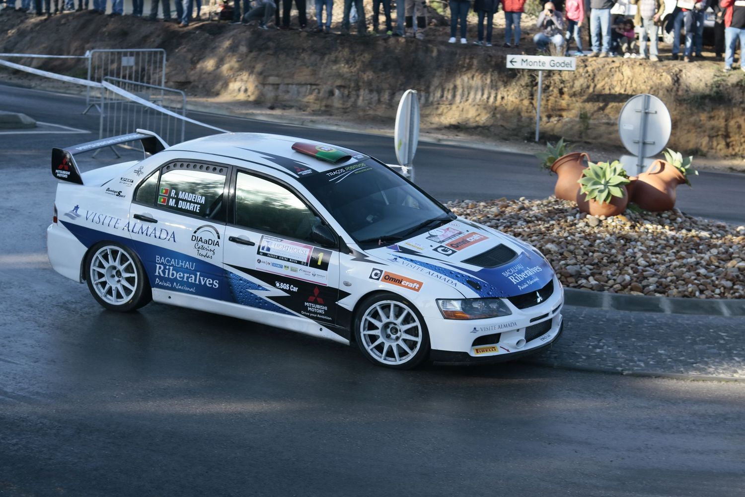 White rally car racing around sharp corner on mountain course with crowd gathered nearby.