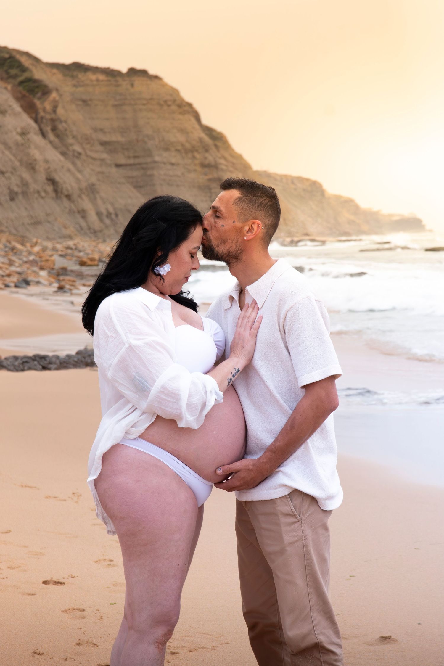 Pregnant couple sharing a tender kiss on a sandy beach at sunset, with rocky cliffs in the background.