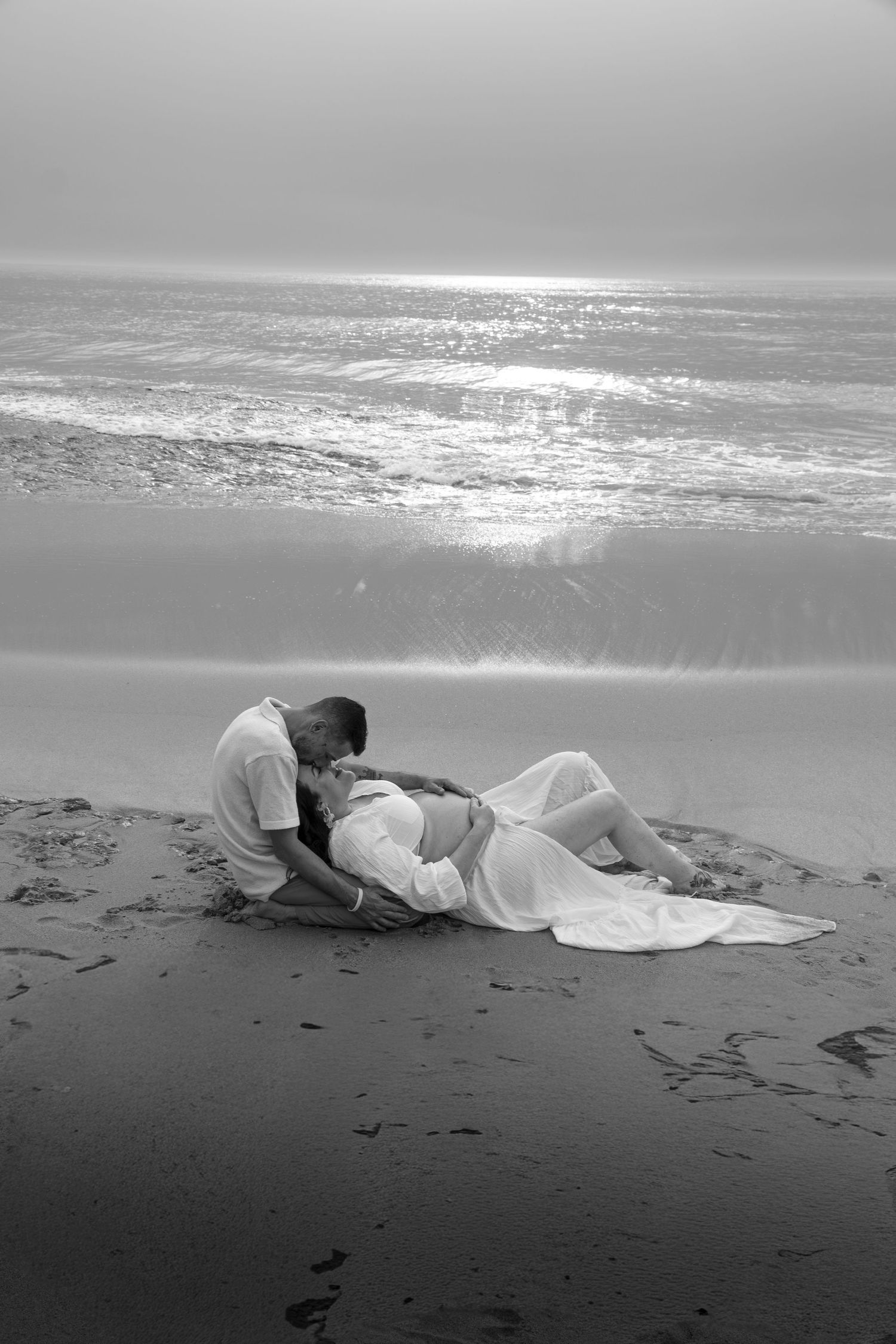 Black and white photo of a romantic couple embracing on a beach with ocean waves reflecting sunlight in the background.