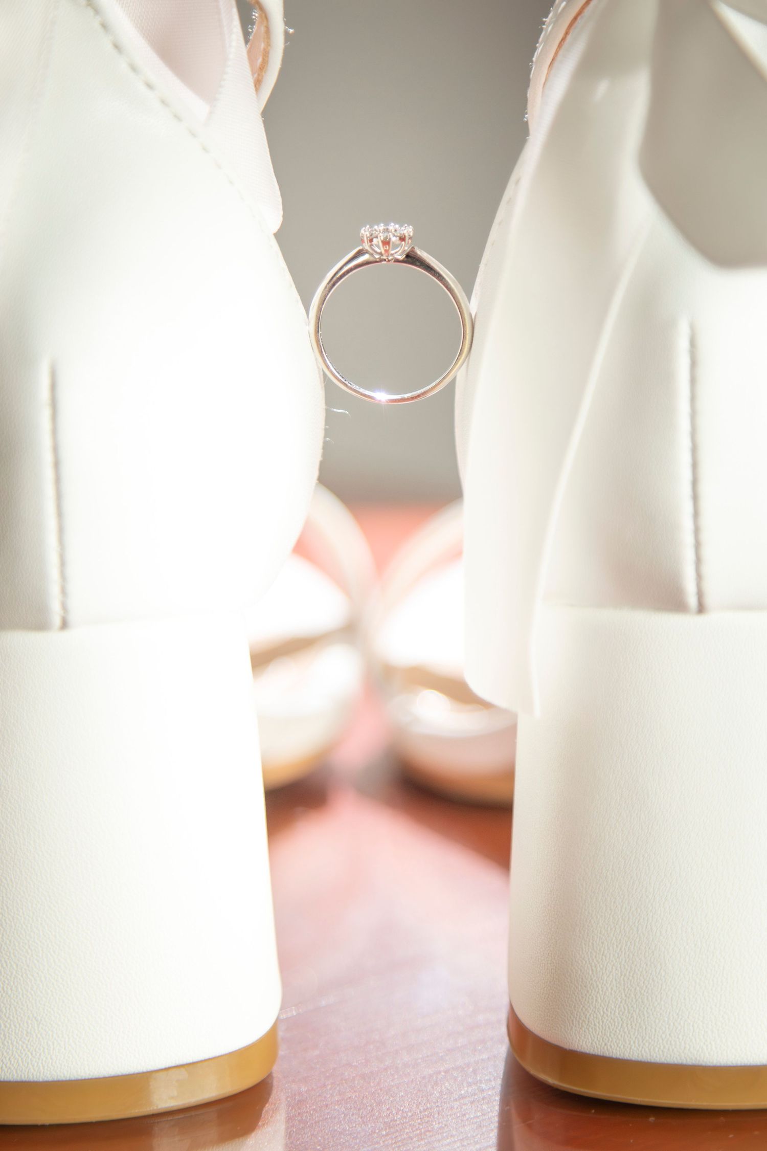 An elegant engagement ring photographed between white wedding shoes.