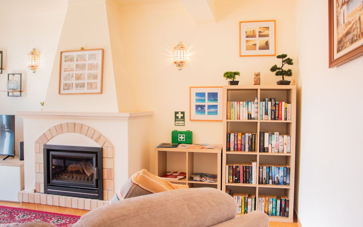 Cozy living room corner with brick fireplace, tall bookshelf, and warm beige walls decorated with framed artwork.