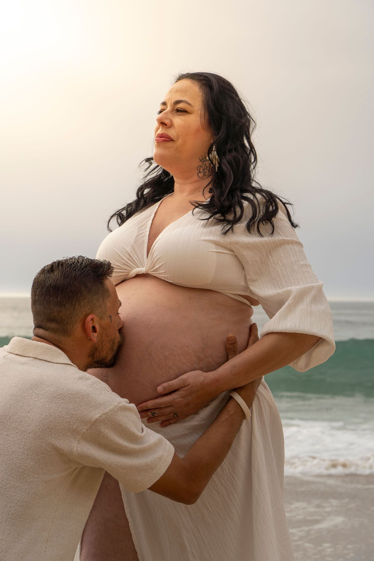 Pregnant woman in white dress at beach with partner kissing her baby bump, maternity photo session by the ocean.