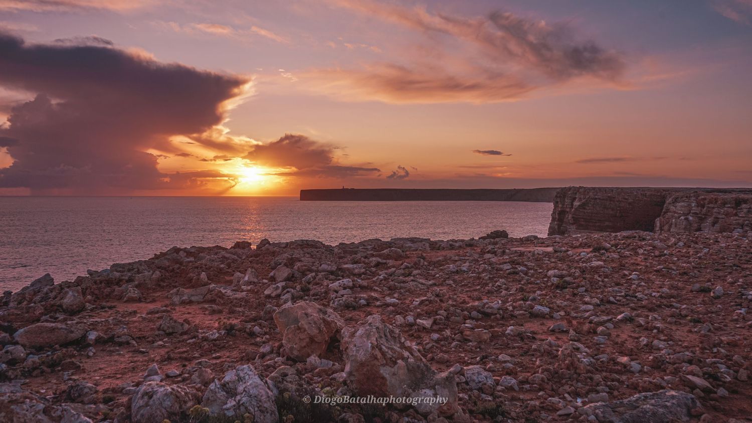Dramatic sunset over rocky coastal cliffs with orange sky reflecting on rugged terrain and ocean waters below.