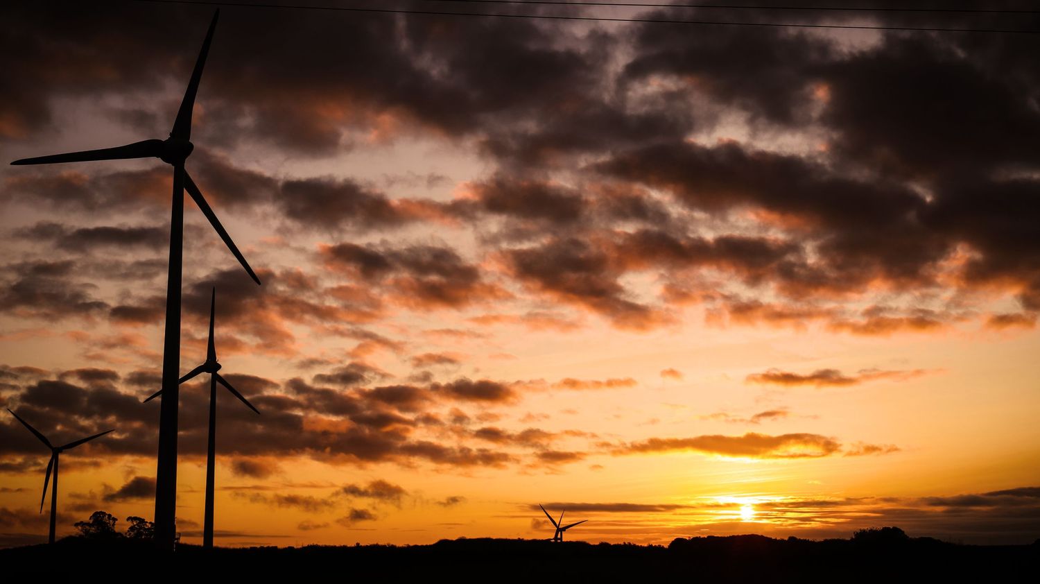 Wind turbines silhouetted against a dramatic orange sunset sky with dark clouds and sun dipping below the horizon.