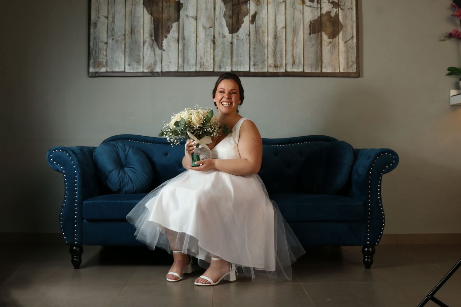 A bride sitting on a blue velvet couch holding flowers beneath a wooden world map wall art.