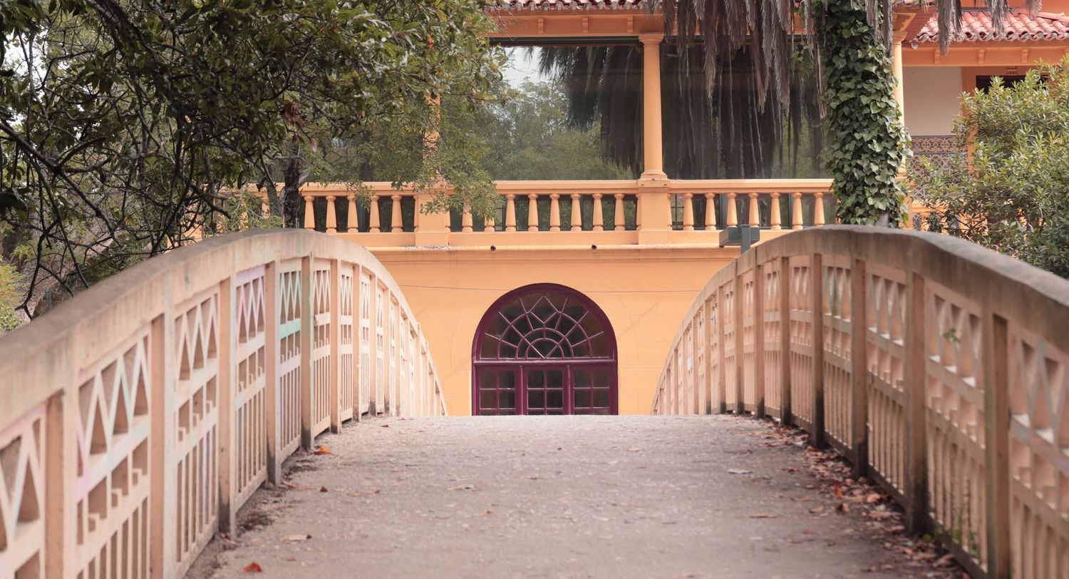 A wooden footbridge leads to an arched doorway entrance framed by pink walls and stone balustrades in a garden setting.