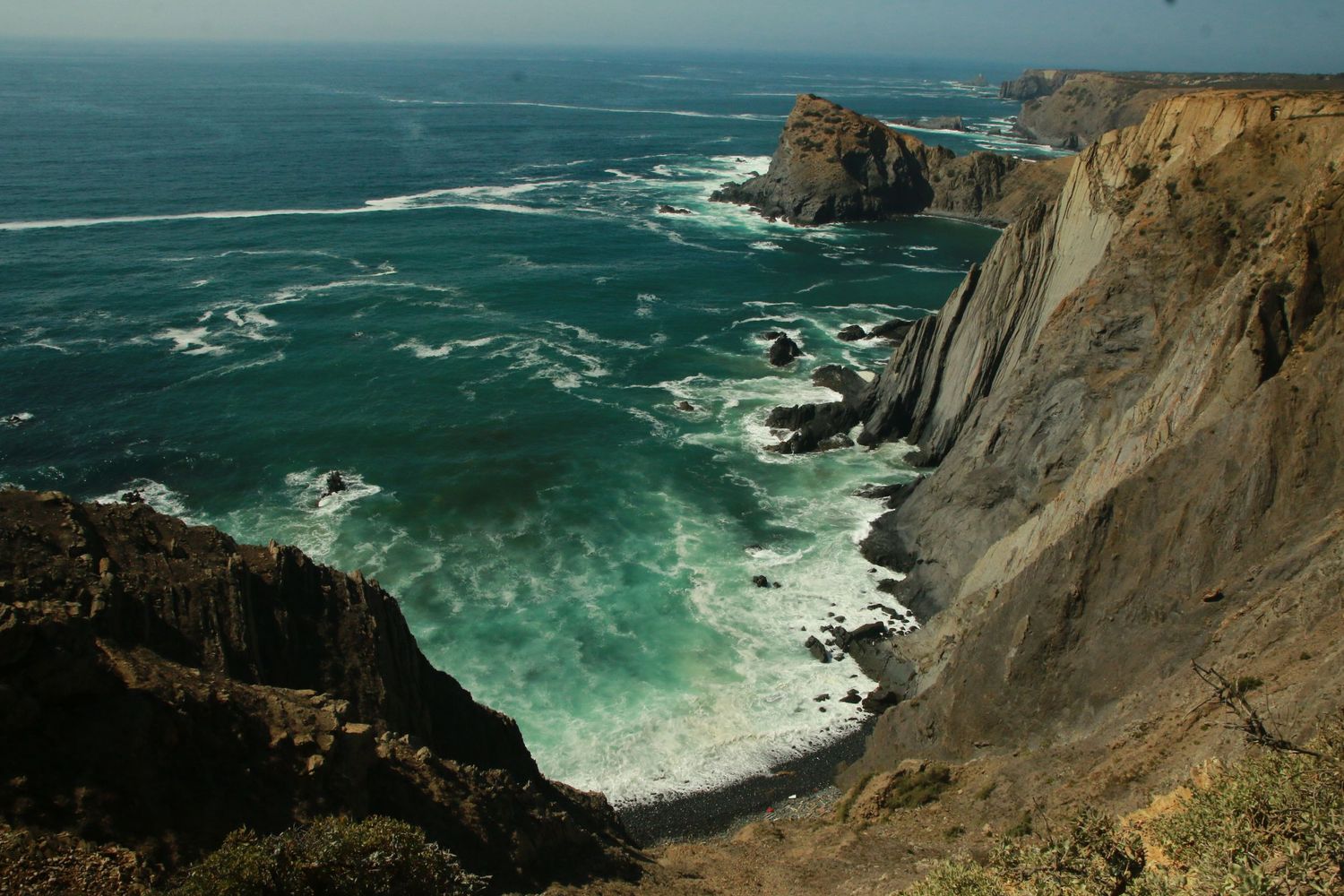 Dramatic coastal cliffs rise above turquoise waves crashing against rocky shores along a rugged California coastline.