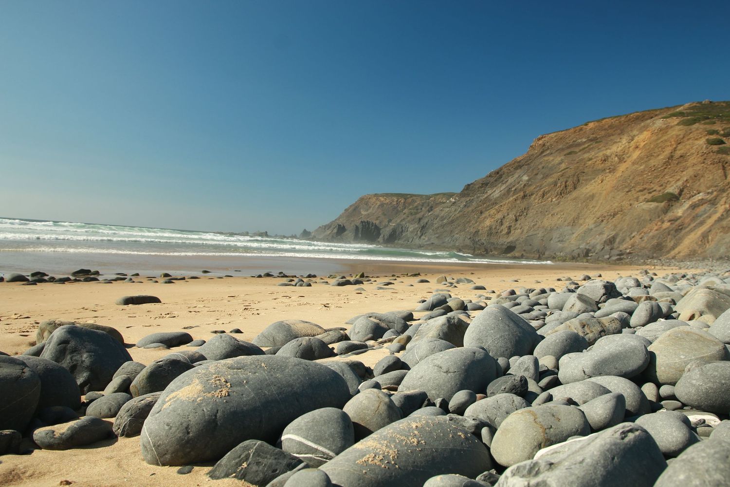 Rocky beach with smooth boulders and coastal cliffs along the Pacific Ocean under clear blue skies.