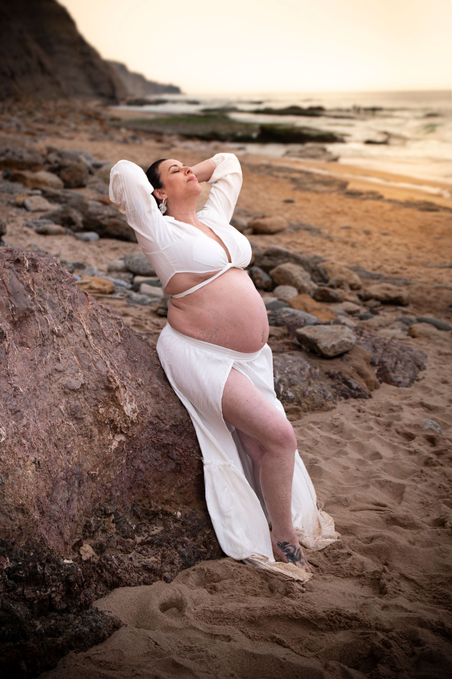 Pregnant woman in white sheer dress posing on sandy beach at sunset, leaning against large rock.