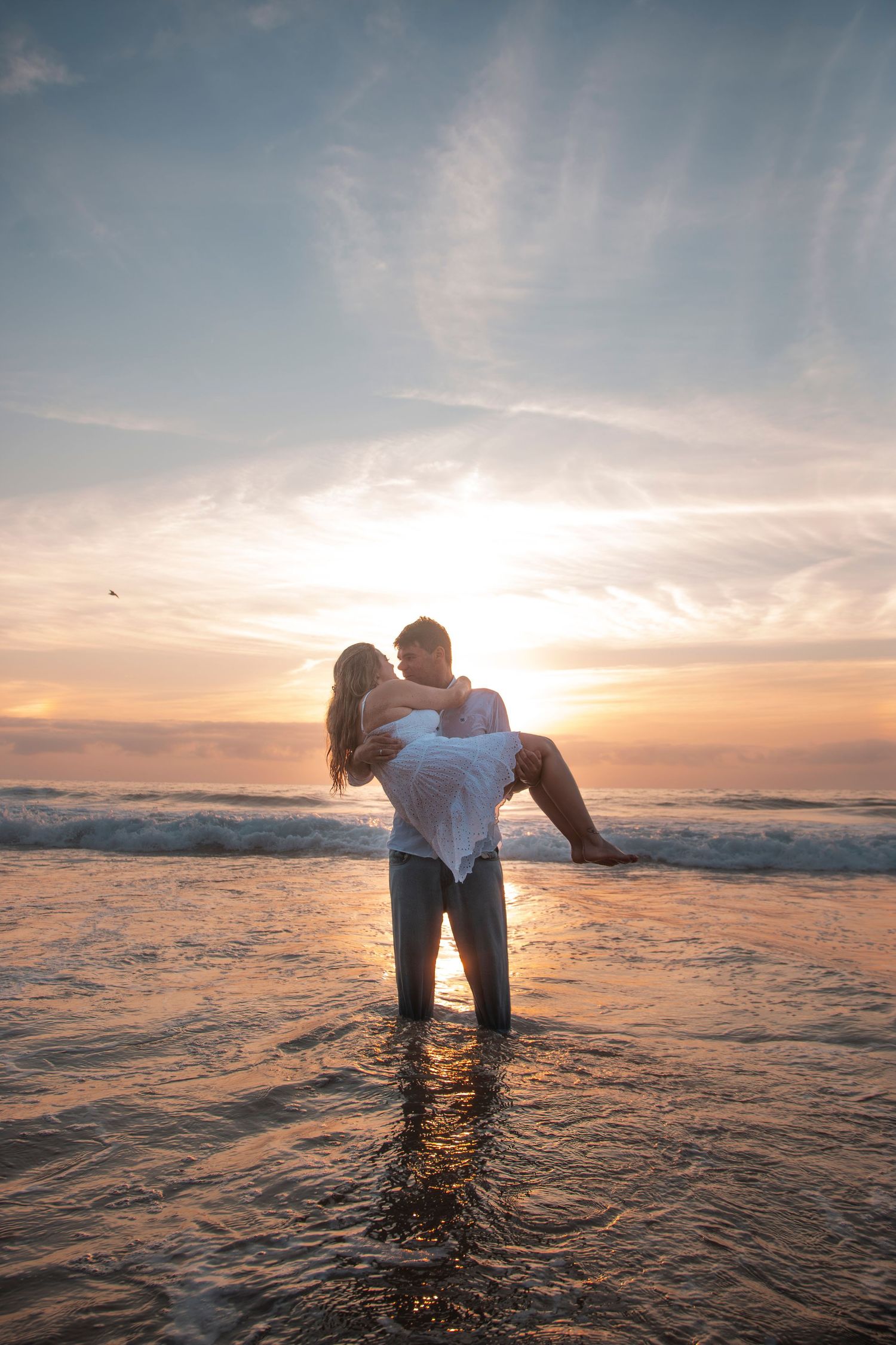 Two silhouettes share a romantic moment while standing in shallow ocean waves during a glowing sunset.