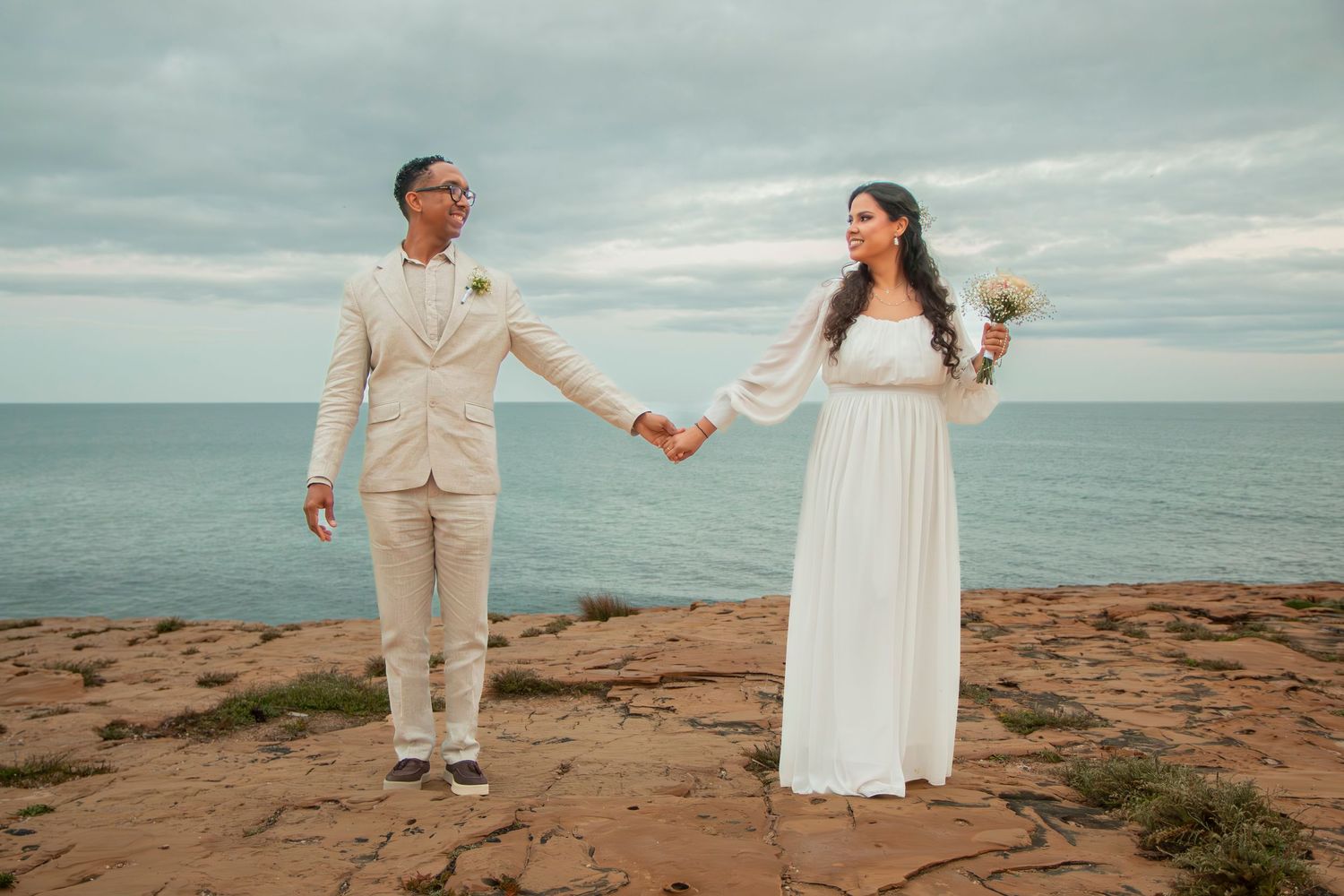 A couple celebrates their wedding day with romantic poses along a scenic rocky ocean coastline at sunset.