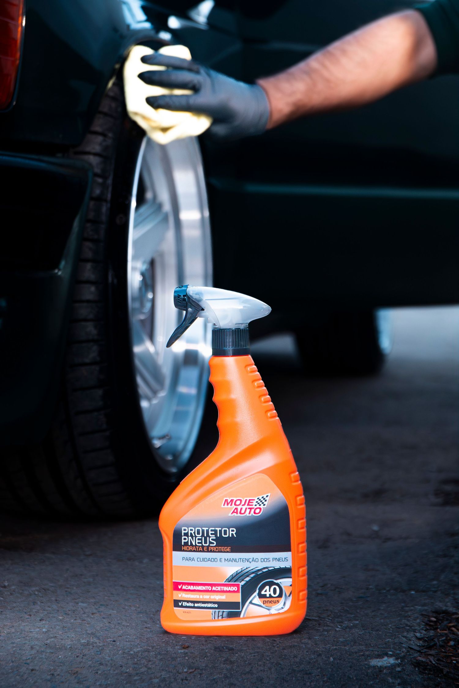 Person cleaning car wheel with cloth and Moje Auto Protetor Pneus tire protector spray bottle in foreground.