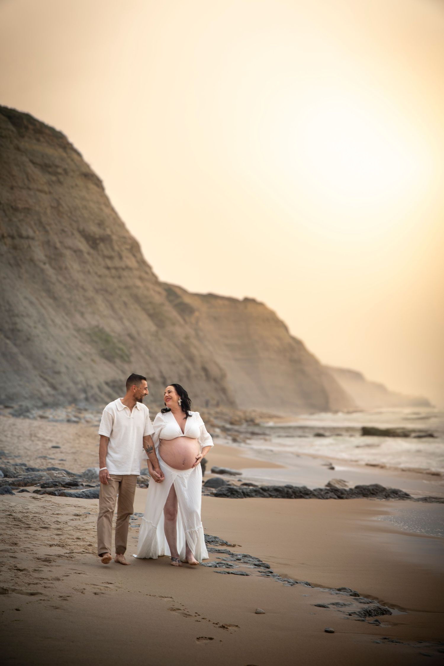 Pregnant couple walks on beach at sunset, woman in white dress cradling baby bump, cliffs in background.