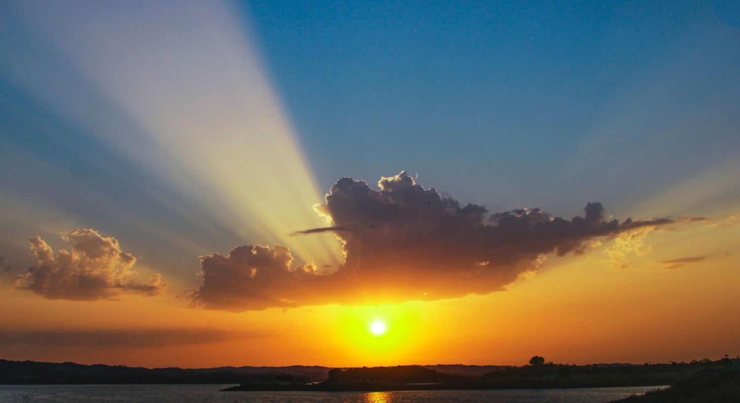 A dramatic sunset over silhouetted landscape with sunbeams breaking through dark clouds in an orange and blue sky.