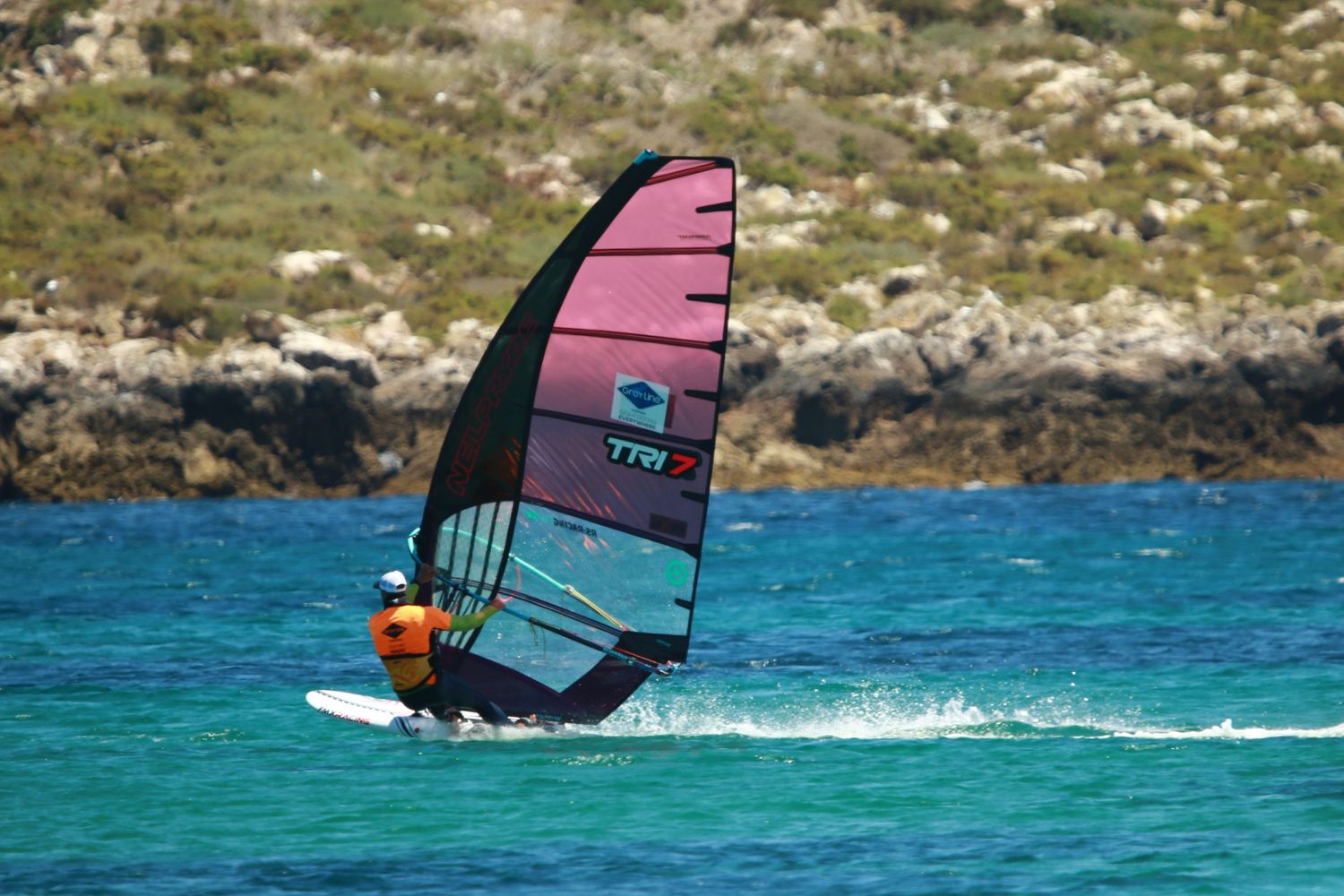 Windsurfer with pink sail gliding through crystal blue waters near rocky shoreline.