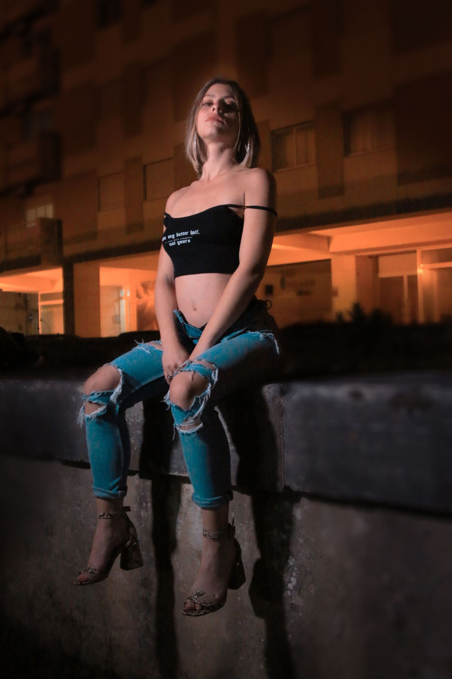 Night portrait showing person in black crop top and distressed blue jeans sitting on concrete ledge with warm lighting.