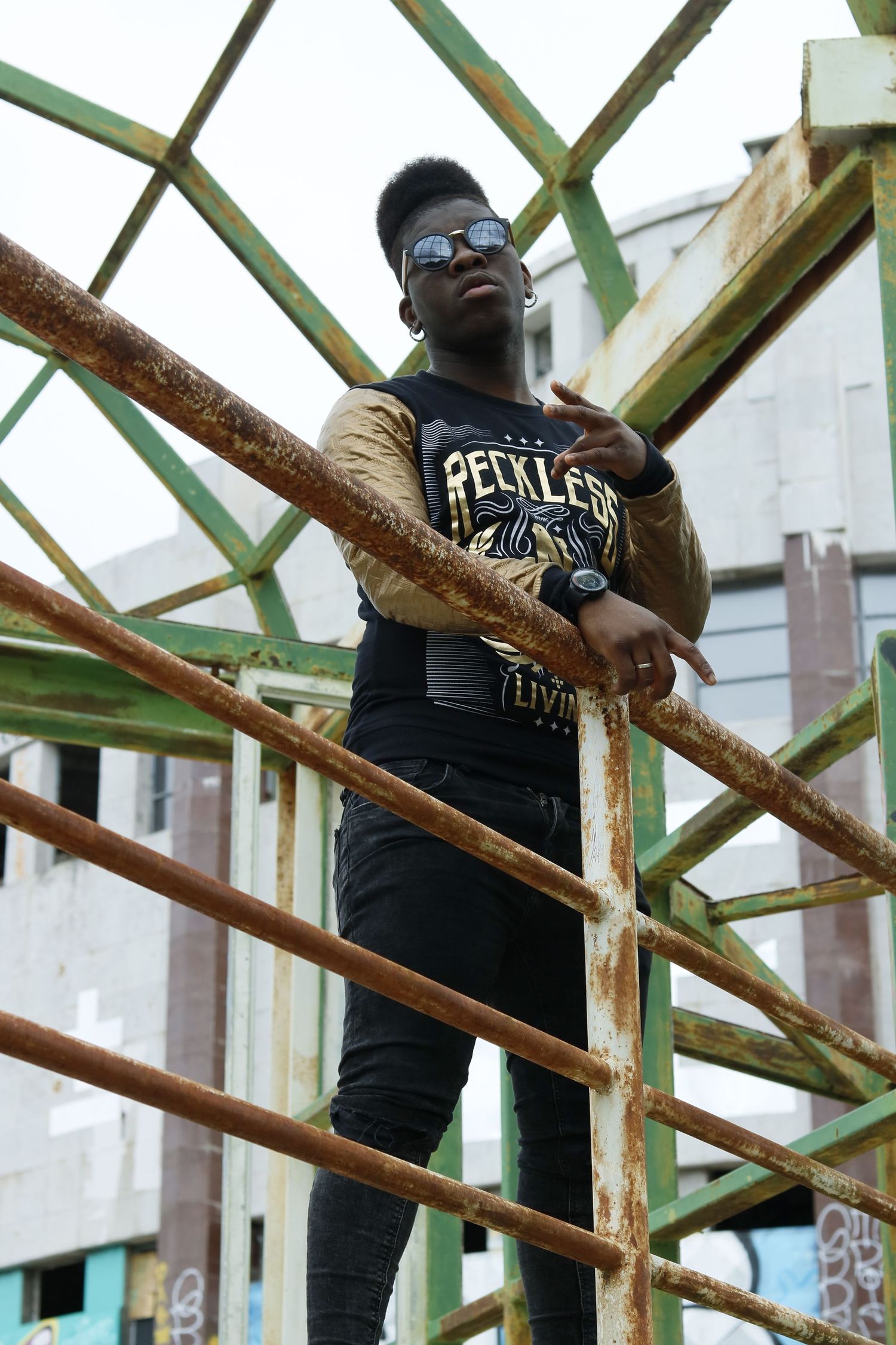 Someone wearing sunglasses and a black t-shirt poses on rusty metal scaffolding against a light colored wall.