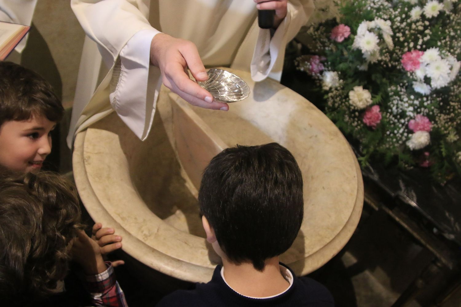 Close-up of baptismal font during religious ceremony with floral decorations visible in background.