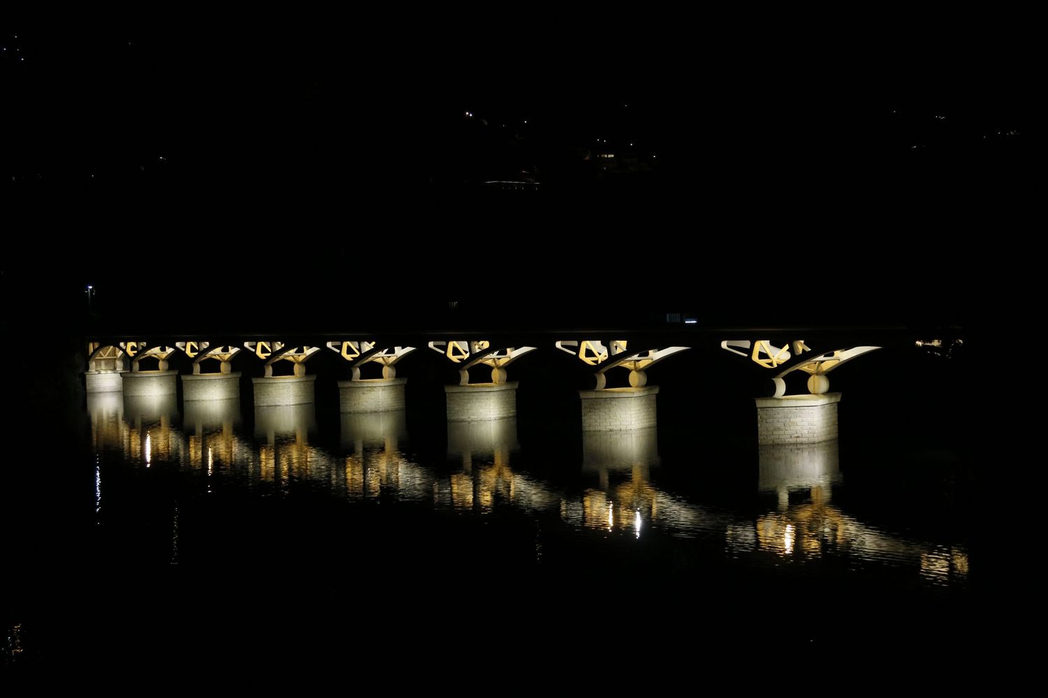 Illuminated concrete bridge pillars reflect off dark water at night creating a symmetrical pattern of lights.