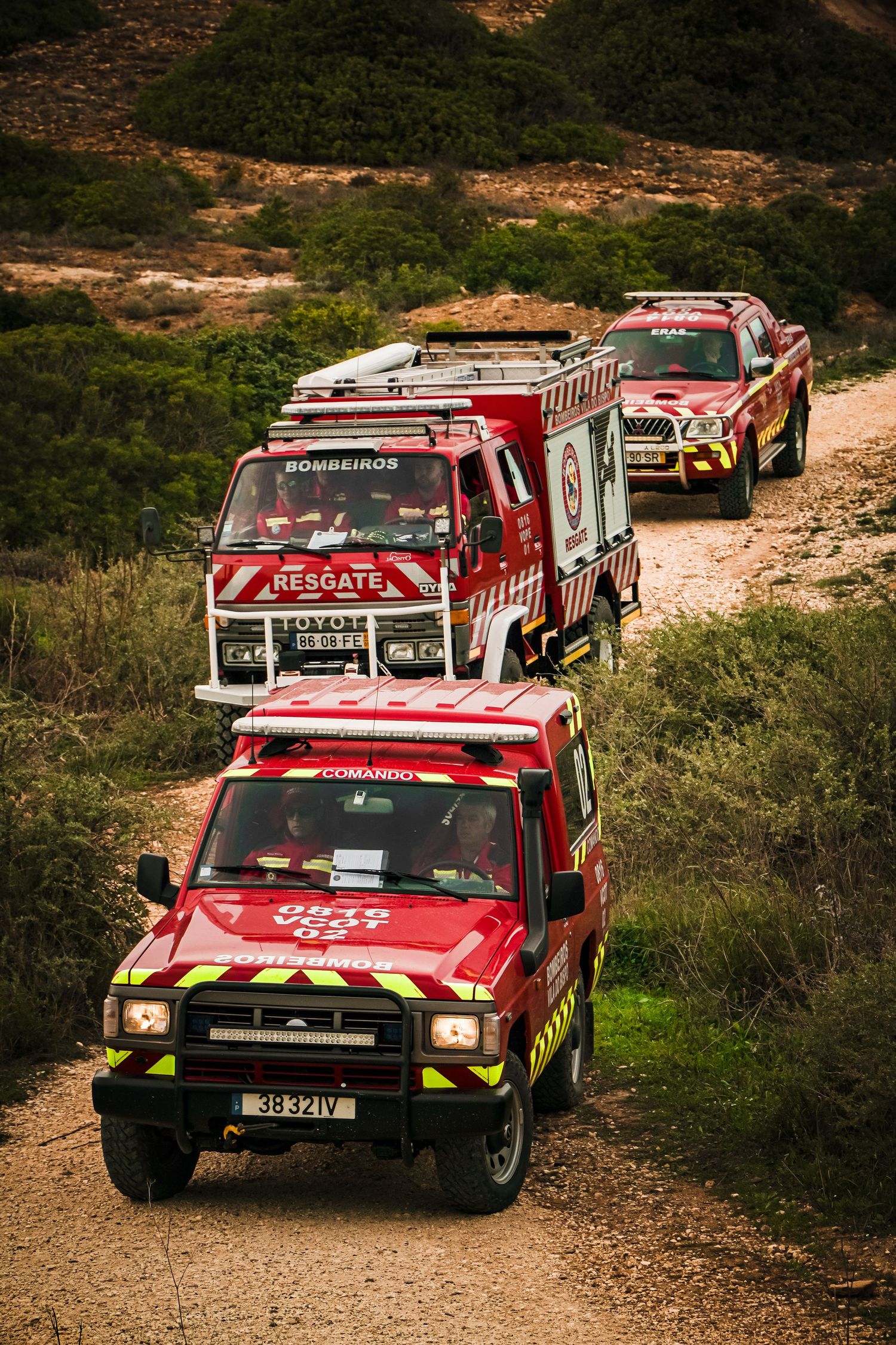 Three red rescue vehicles driving in formation along a dirt mountain road.