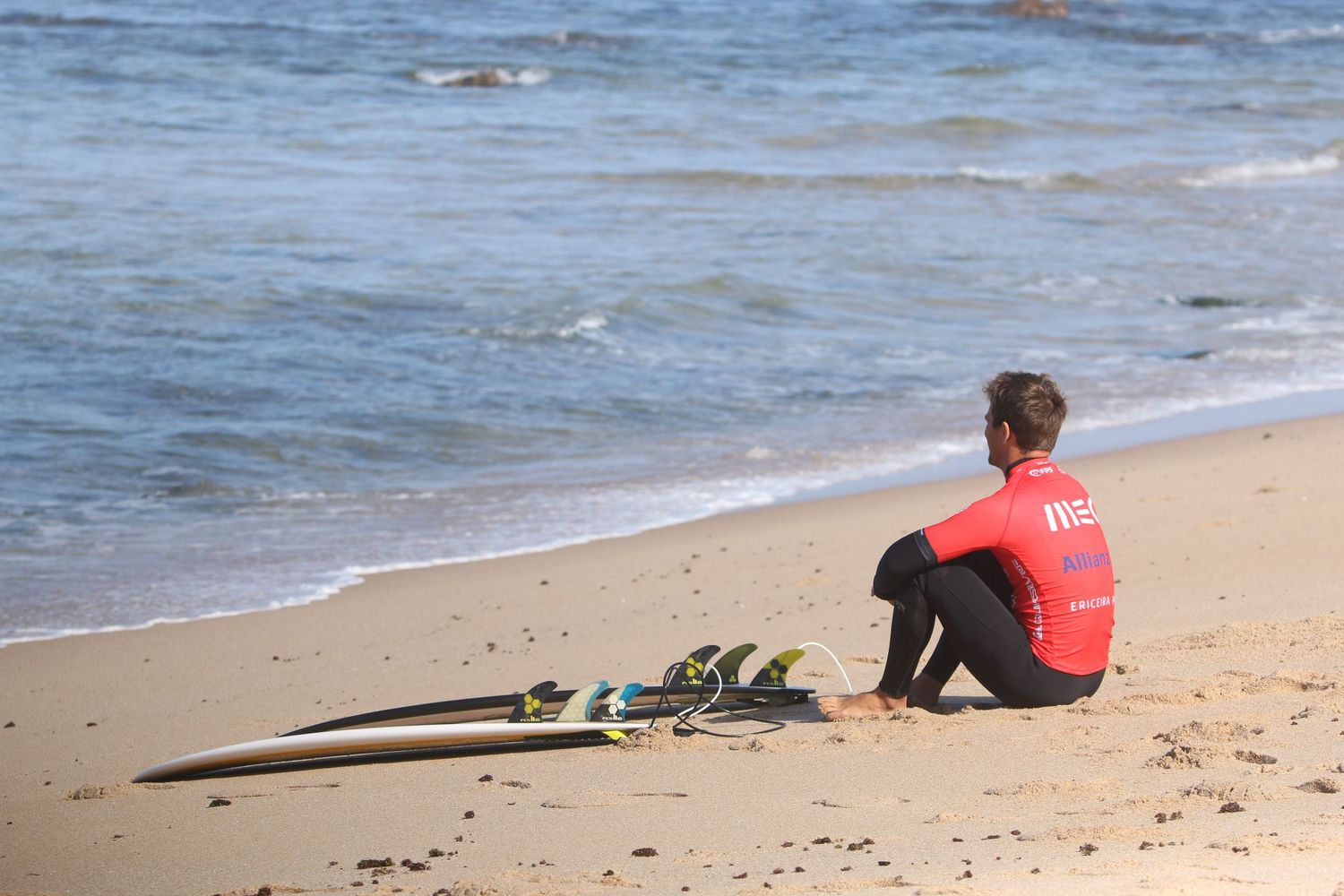 Person in red wetsuit sitting on sandy beach next to surfboard while watching ocean waves on sunny day.