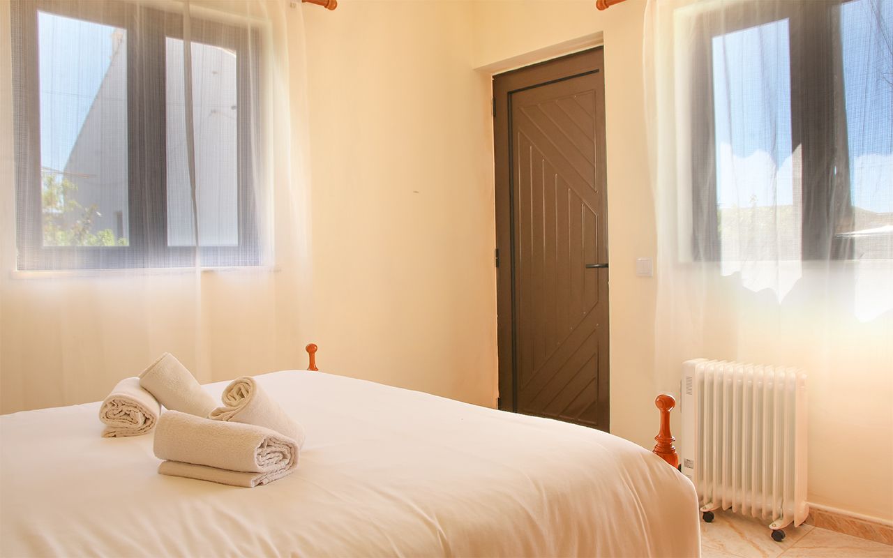 Bright bedroom interior with white bedding, windows on both sides, dark wooden door and white radiator beneath window.