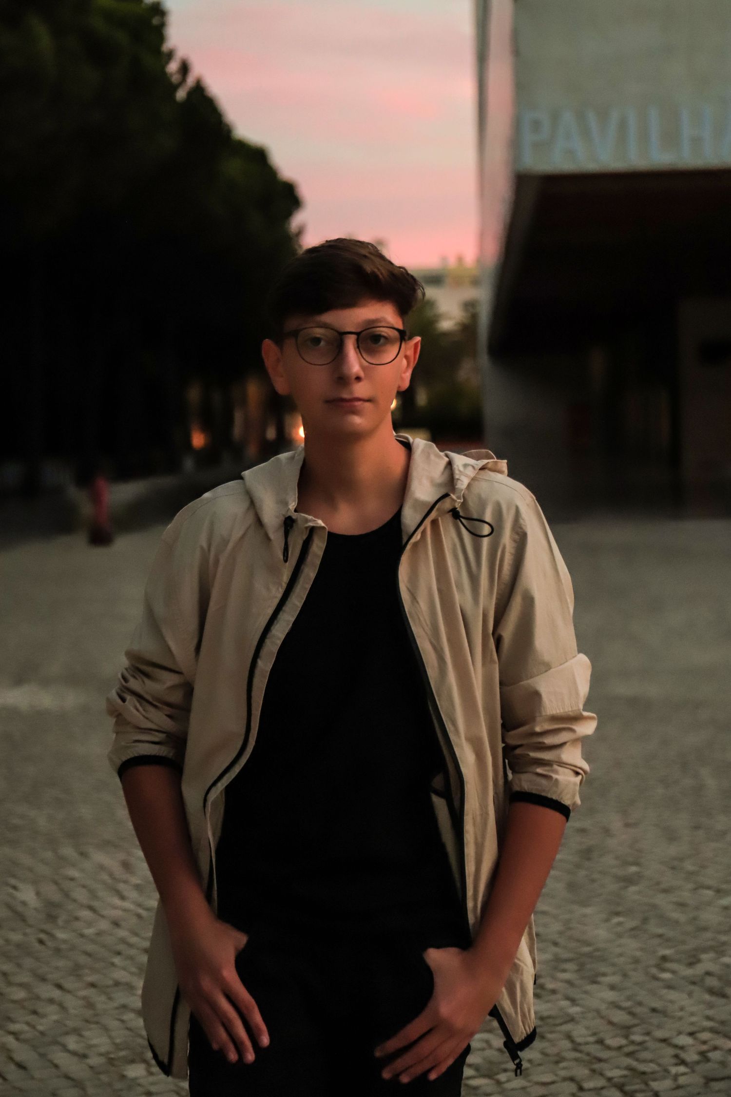 Figure in beige jacket and glasses standing outdoors during sunset near a pavilion.
