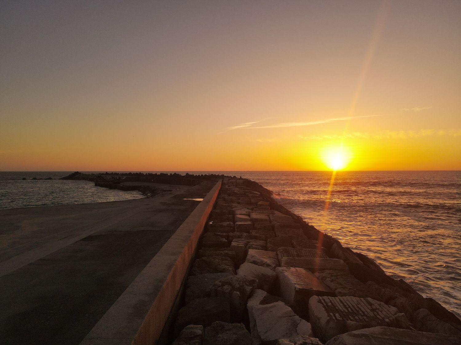 Dramatic sunset over ocean with sun rays illuminating rocky jetty breakwater extending into calm waters.