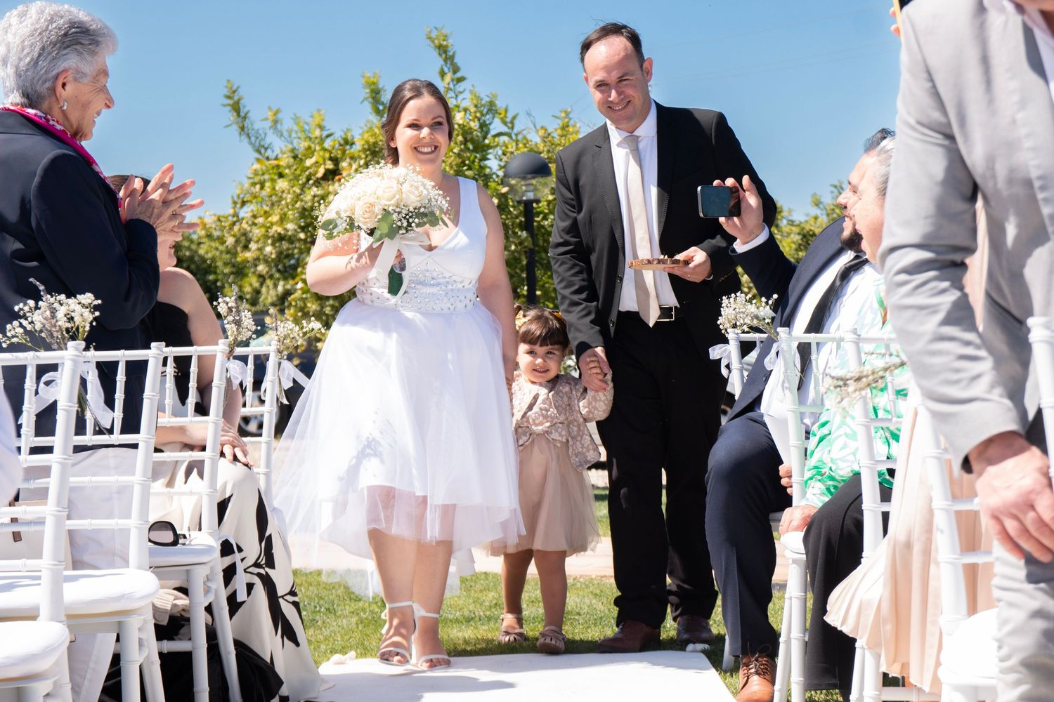 Wedding ceremony procession on outdoor patio with white chairs and bright sunny sky.