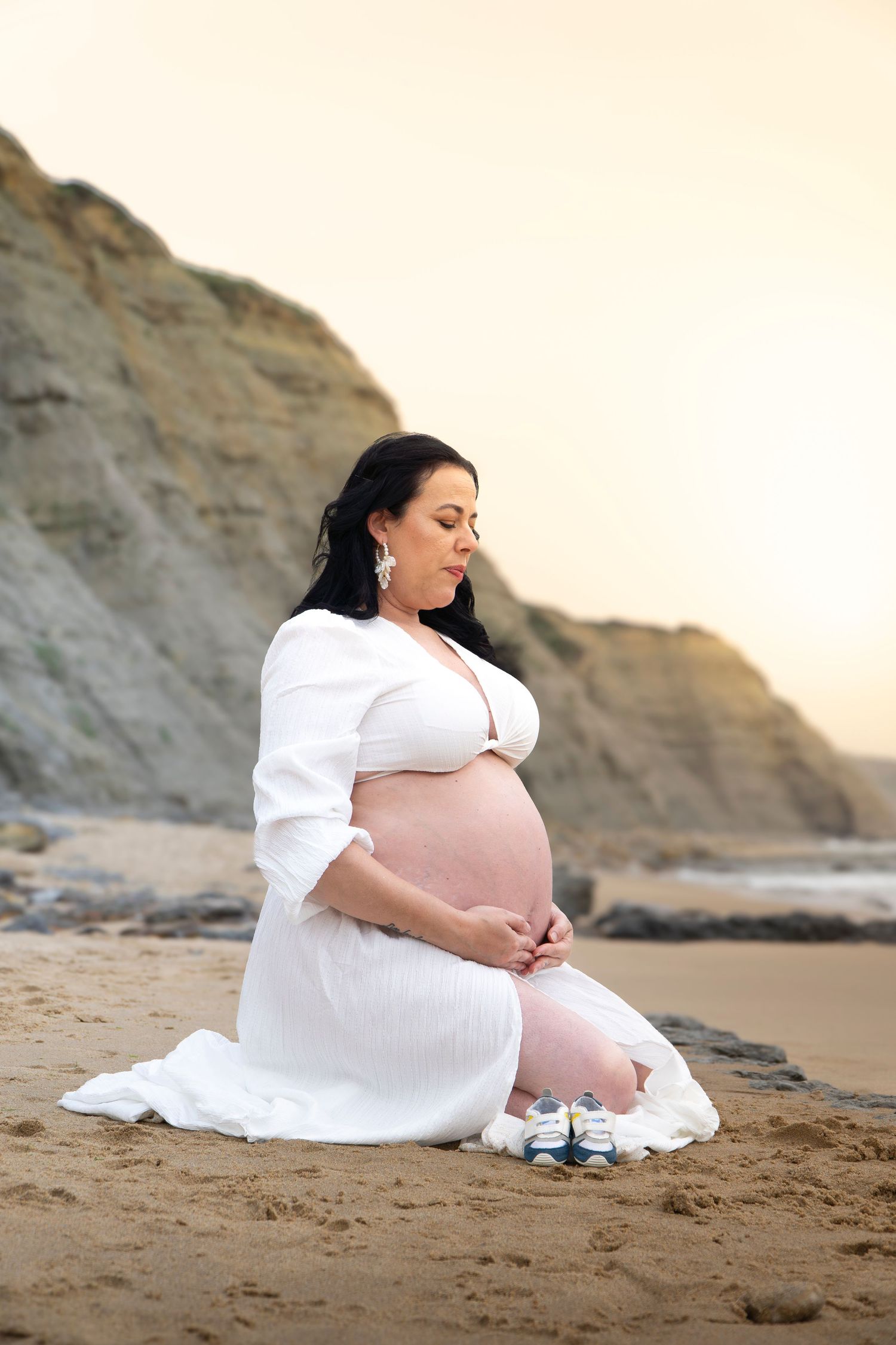 Pregnant woman in white dress sitting on sandy beach, cradling her belly near rocky cliffs at sunset.