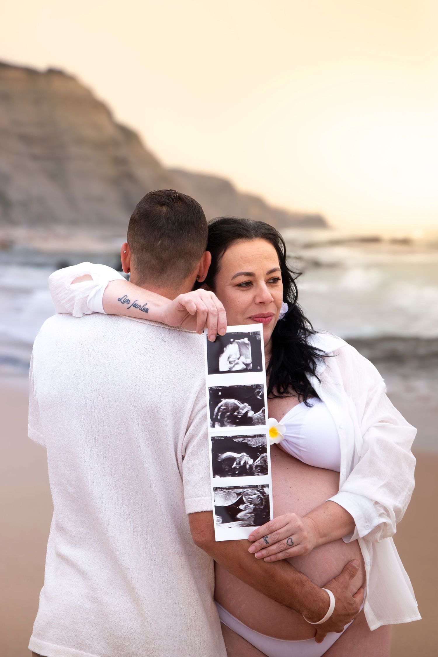 Pregnant couple embracing on beach at sunset, holding ultrasound photos, woman in white bikini and shirt.