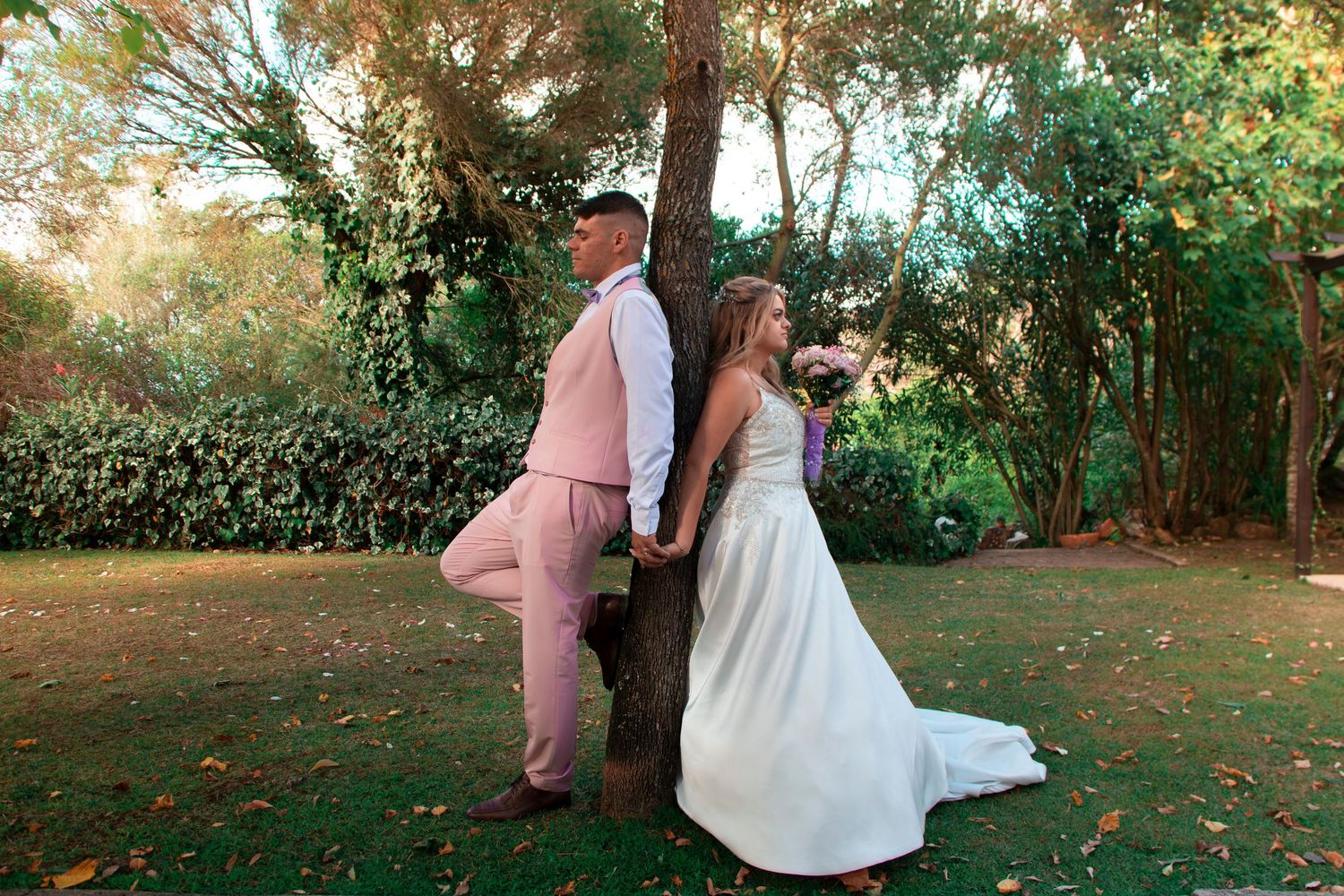 Bride and groom in pink suit stand back-to-back against a tree in a lush garden on their wedding day.