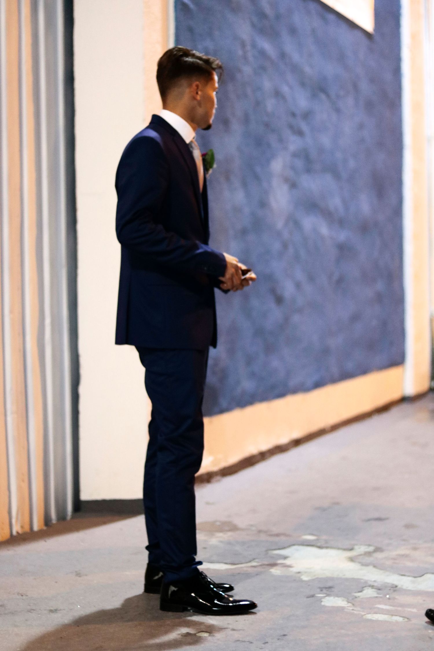 A groom in a navy blue suit and polished shoes stands in profile against a textured wall.