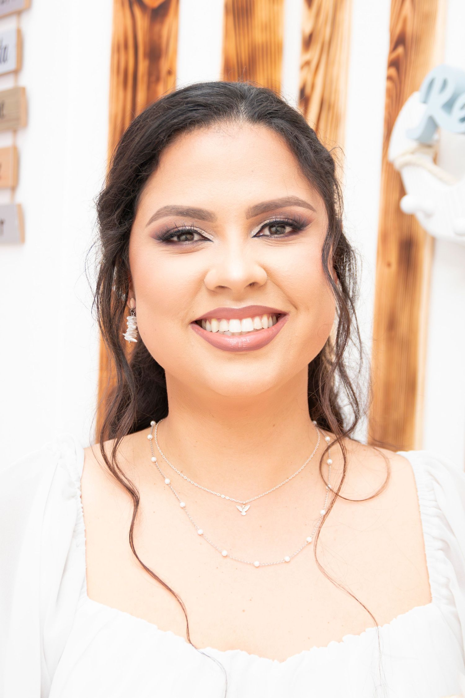 A smiling woman wearing a white top with delicate necklace and dark eye makeup in front of curtained window.