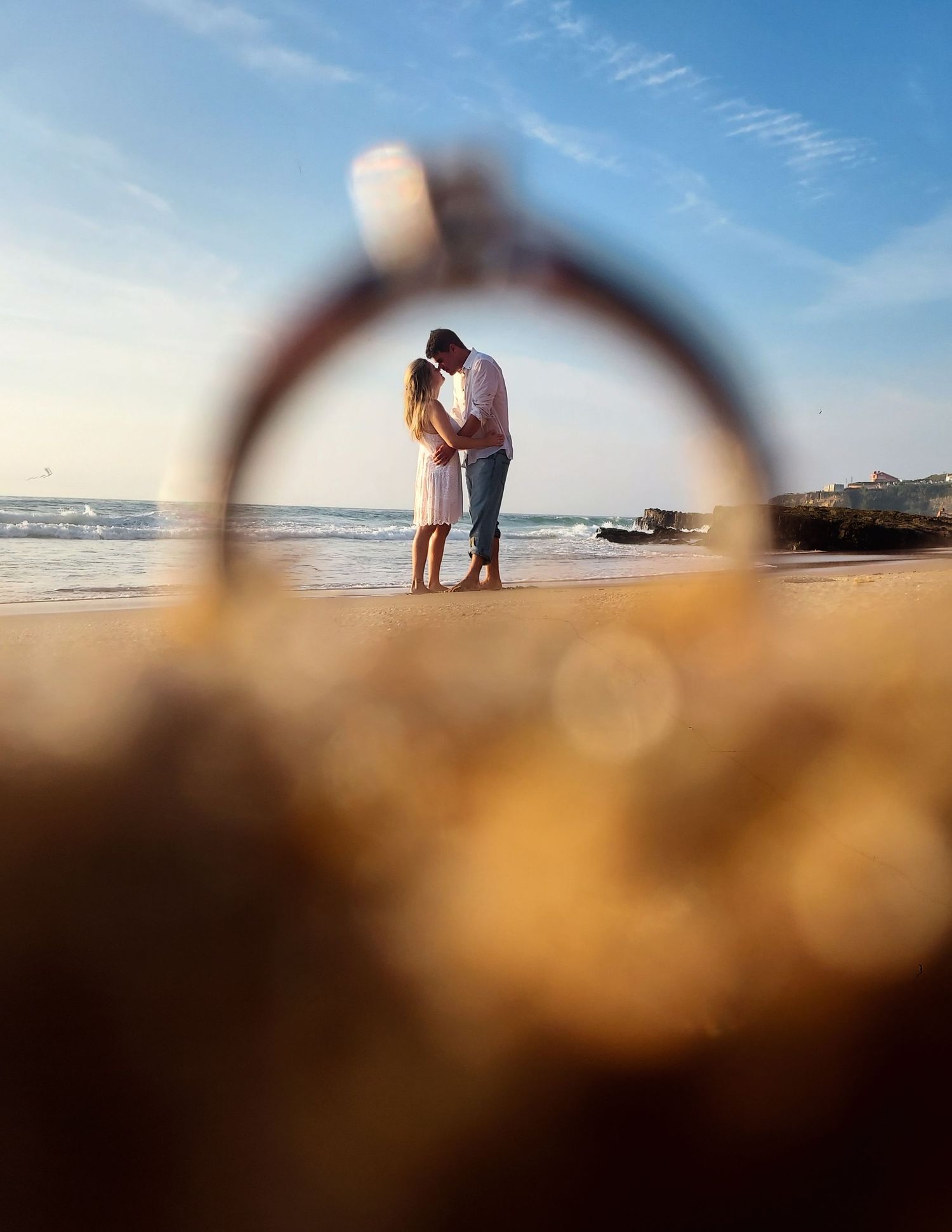 A romantic couple silhouetted on a beach is viewed through a golden ring at sunset.