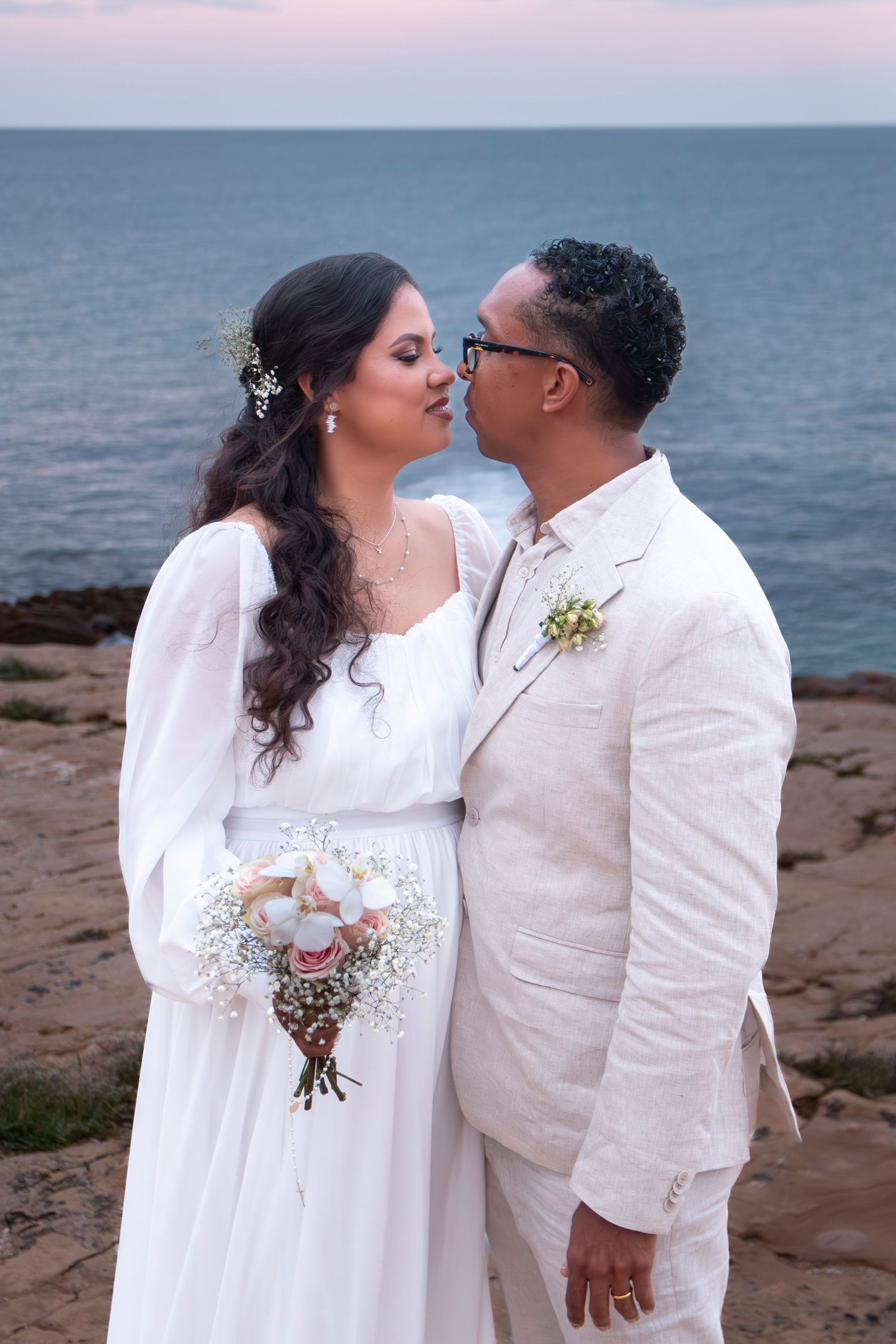 A romantic wedding couple shares intimate moments on a beach at sunset, dressed in matching white outfits.