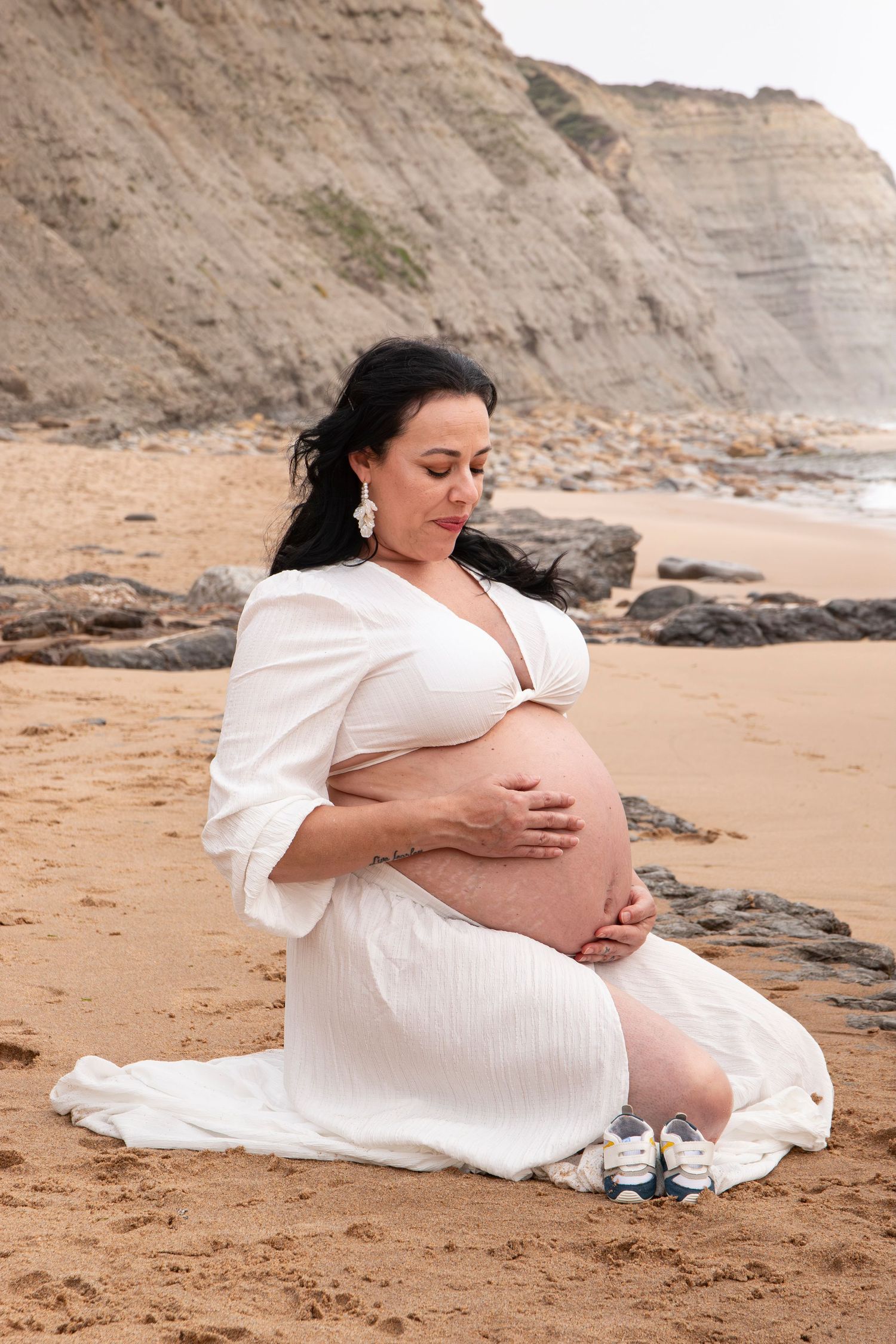 Pregnant woman in white dress sitting on sandy beach, cradling her baby bump with rocky cliffs in background.