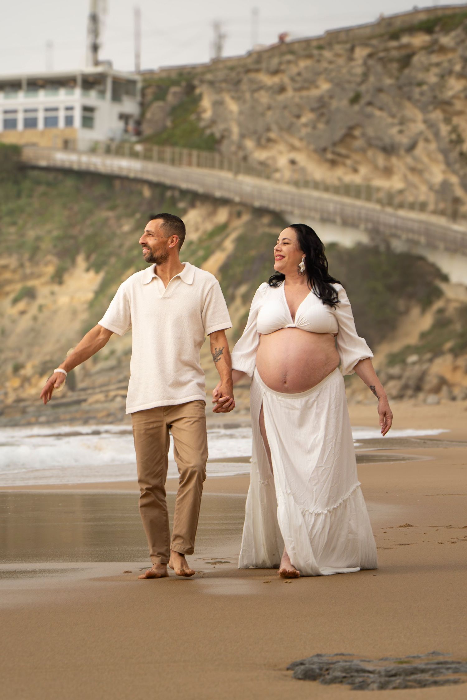 Pregnant woman in white dress walks hand-in-hand with partner on sandy beach near rocky cliffs.