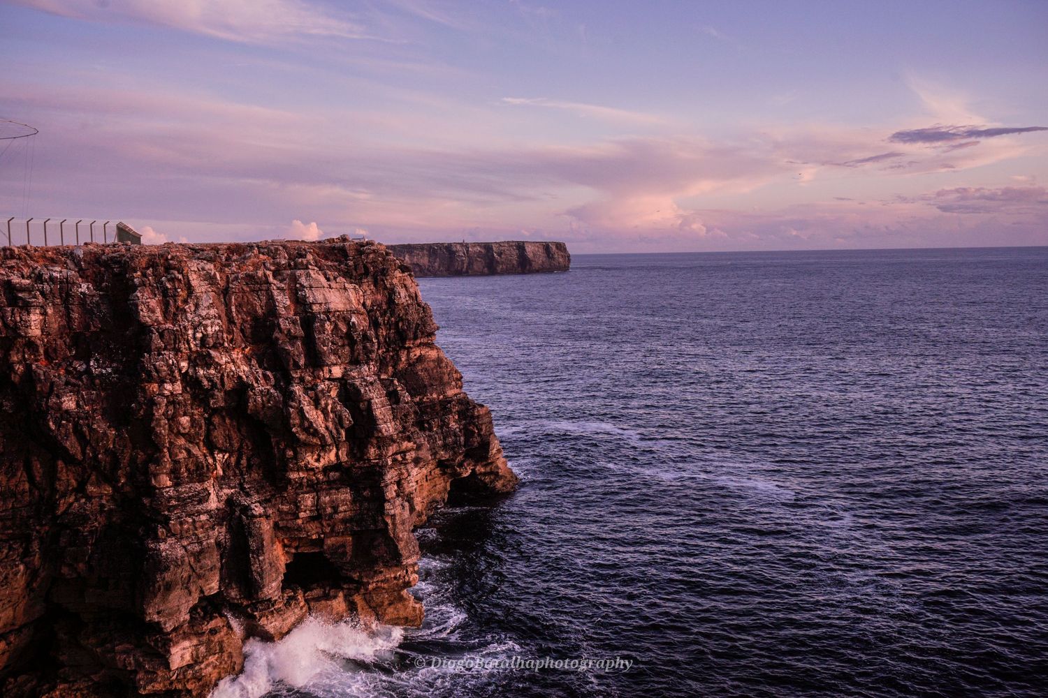Dramatic rocky cliffs tower over the Atlantic Ocean at sunset with pink and purple sky in Sagres, Portugal.