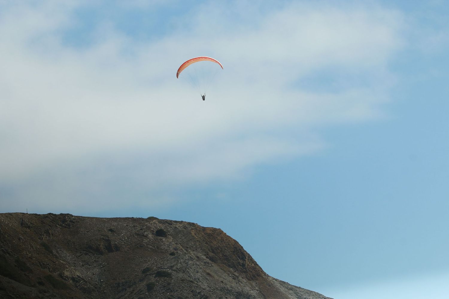 Orange paraglider soaring against a blue sky over a rocky mountain peak.