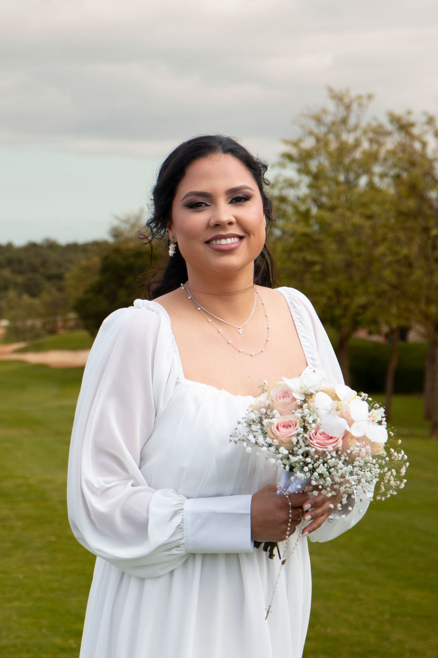 A bride in a white long-sleeved dress holds a delicate bouquet of pink roses and baby's breath in an outdoor garden setting.