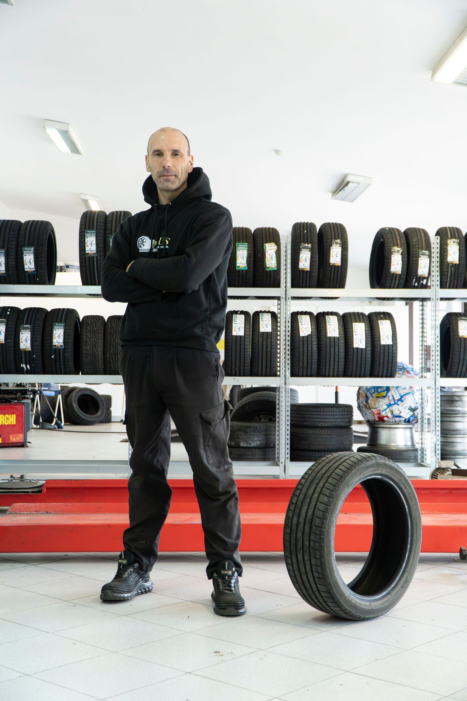 Bald mechanic in black hoodie stands arms crossed beside a tire in a well-stocked tire shop with shelved tires behind him.