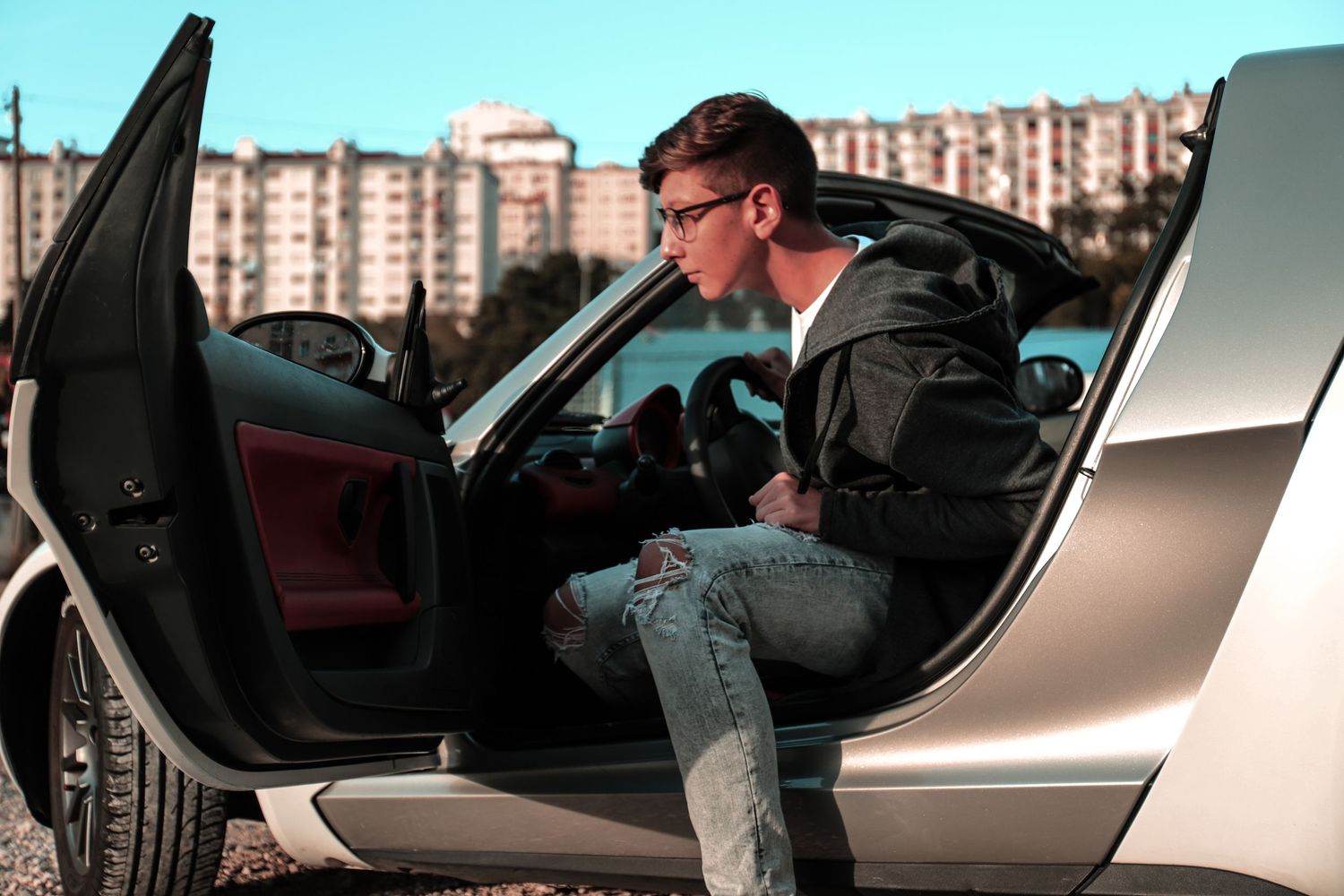 A person in sunglasses gets into a silver car wearing ripped jeans and a black jacket in an urban setting.