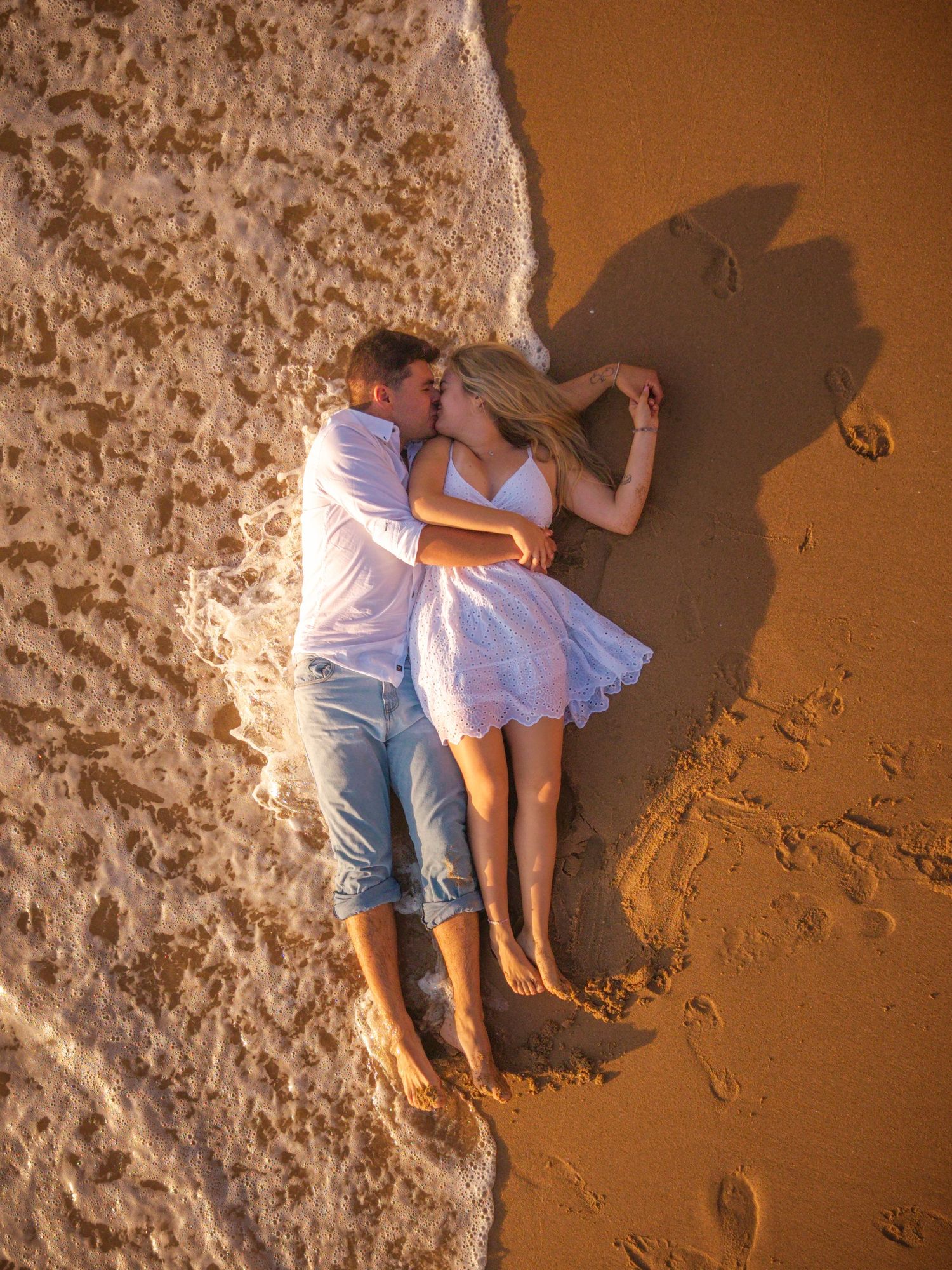A couple in white and blue outfits laying on sandy beach creating shadows at sunset on the shoreline.
