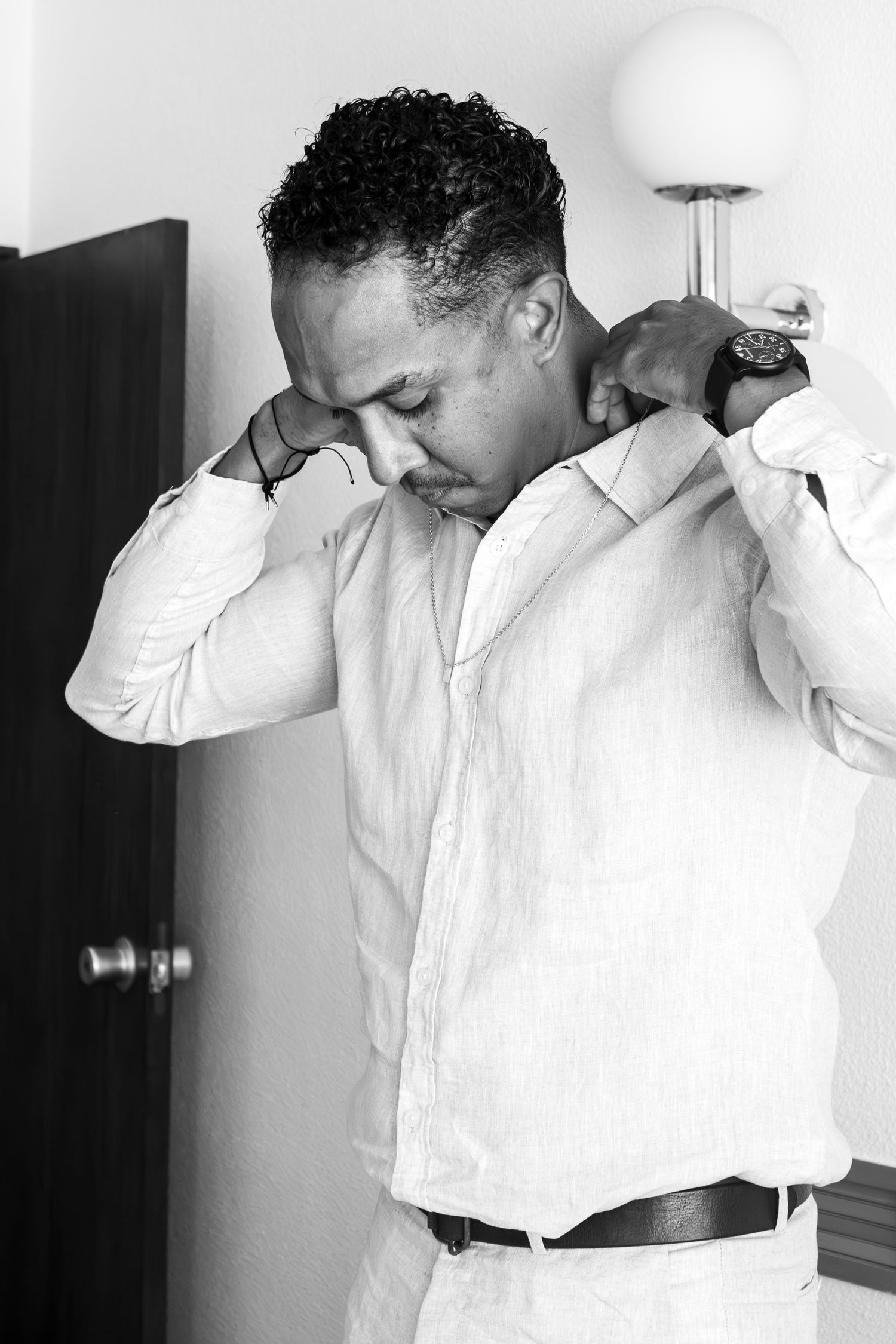 A person wearing a white button-up shirt is getting dressed in a room with a lamp in the background.